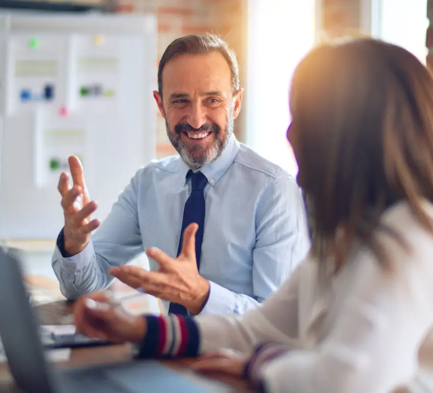 A smiling businessman in a blue shirt and tie having a discussion with a colleague in an office with charts visible in the background.
